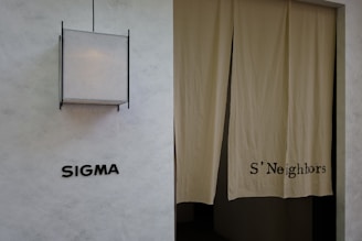 A minimalist exterior view featuring a textured wall with the word 'SIGMA' displayed in bold black letters. To the right, a pair of beige curtains partly covers a doorway, with 'S'Neighbors' printed on the fabric. A geometric light fixture with a square design is installed above the signage.