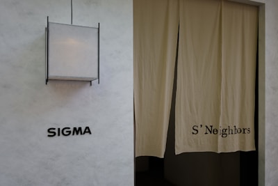 A minimalist exterior view featuring a textured wall with the word 'SIGMA' displayed in bold black letters. To the right, a pair of beige curtains partly covers a doorway, with 'S'Neighbors' printed on the fabric. A geometric light fixture with a square design is installed above the signage.