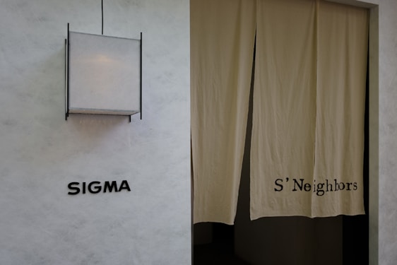 A minimalist exterior view featuring a textured wall with the word 'SIGMA' displayed in bold black letters. To the right, a pair of beige curtains partly covers a doorway, with 'S'Neighbors' printed on the fabric. A geometric light fixture with a square design is installed above the signage.