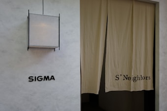 A minimalist exterior view featuring a textured wall with the word 'SIGMA' displayed in bold black letters. To the right, a pair of beige curtains partly covers a doorway, with 'S'Neighbors' printed on the fabric. A geometric light fixture with a square design is installed above the signage.