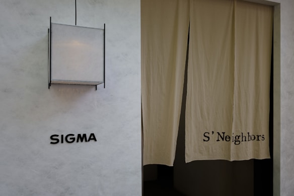 A minimalist exterior view featuring a textured wall with the word 'SIGMA' displayed in bold black letters. To the right, a pair of beige curtains partly covers a doorway, with 'S'Neighbors' printed on the fabric. A geometric light fixture with a square design is installed above the signage.