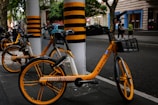 Three bicycles with bright yellow and black accents are parked on a sidewalk near tall poles painted with matching yellow and black stripes. The scene takes place on a street in an urban environment, with a few pedestrians visible in the background.