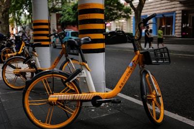 Three bicycles with bright yellow and black accents are parked on a sidewalk near tall poles painted with matching yellow and black stripes. The scene takes place on a street in an urban environment, with a few pedestrians visible in the background.