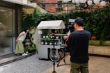 A person operates a camera setup, recording or photographing a scene outdoors. Nearby, another person in a white t-shirt and green skirt leans forward to look closely at a small cart filled with potted plants. This scene is set against a backdrop of a building and a lush, ivy-covered wall, creating a contrast between modern elements and nature. Several paper lanterns hang overhead, contributing to the ambient decor.