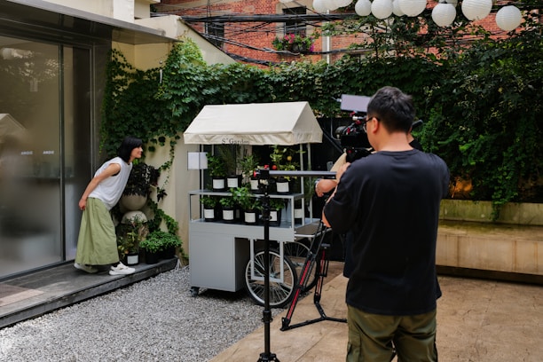 A person operates a camera setup, recording or photographing a scene outdoors. Nearby, another person in a white t-shirt and green skirt leans forward to look closely at a small cart filled with potted plants. This scene is set against a backdrop of a building and a lush, ivy-covered wall, creating a contrast between modern elements and nature. Several paper lanterns hang overhead, contributing to the ambient decor.
