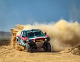 A racing truck is speeding along a dirt track, kicking up a large cloud of dust behind it. The truck has green and red colored bodywork with various decals and the name 'Team Sultan' prominently displayed. The terrain is arid and dusty, with a clear blue sky overhead.