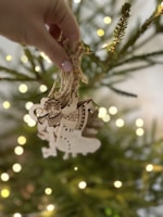 A hand holds a bundle of wooden ornaments in front of a blurred, decorated Christmas tree. The ornaments feature intricate designs, including musical notes and a festive figure playing a trumpet. Warm fairy lights softly illuminate the background, adding a cozy glow.