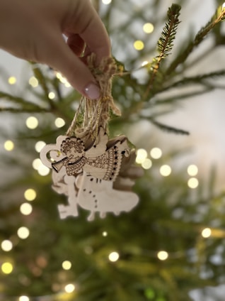 A hand holds a bundle of wooden ornaments in front of a blurred, decorated Christmas tree. The ornaments feature intricate designs, including musical notes and a festive figure playing a trumpet. Warm fairy lights softly illuminate the background, adding a cozy glow.