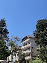 A multi-story residential building with balconies is surrounded by tall, leafy trees. The sky is clear and blue, and power lines crisscross the upper part of the scene. Some signage is visible near the base of the building, indicating a commercial area at street level.