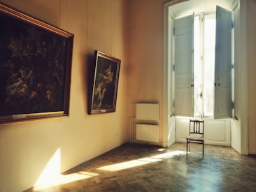 A serene room bathes in soft natural light streaming through partially open shutters. The wooden floor reflects the warm tones from the sunlight. On the left wall, two dark, classical paintings with ornate frames hang at an angle, casting slight shadows. A solitary wooden chair is placed near the window, contributing to a contemplative atmosphere.