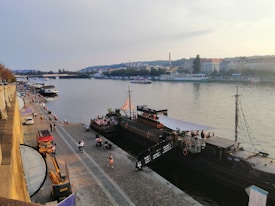 A scenic river scene featuring a paved walkway along the waterfront with people walking and interacting. Multiple ships, including a large boat styled like a pirate ship, are docked by the river. The background features a cityscape with buildings and trees under a partly cloudy sky.