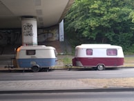 Two vintage caravans are parked under a bridge with graffiti on the support pillars. One caravan is blue and white, and the other is maroon and white. The area around is paved and surrounded by greenery.