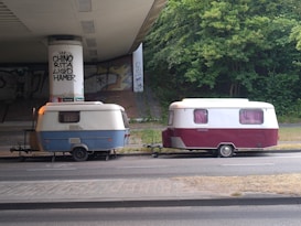 Two vintage caravans are parked under a bridge with graffiti on the support pillars. One caravan is blue and white, and the other is maroon and white. The area around is paved and surrounded by greenery.