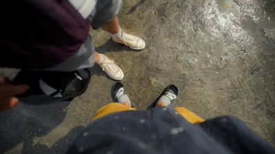 Two pairs of feet are visible on a textured, wet surface. One person is wearing light-colored canvas sneakers, while the other is wearing black sandals with socks. The clothing visible includes shorts and a jacket or sweater. The image provides a casual perspective, as if looking down at one's own feet while standing next to someone.