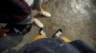 Two pairs of feet are visible on a textured, wet surface. One person is wearing light-colored canvas sneakers, while the other is wearing black sandals with socks. The clothing visible includes shorts and a jacket or sweater. The image provides a casual perspective, as if looking down at one's own feet while standing next to someone.