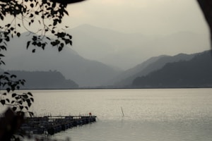 A serene lakeside view with misty, layered mountains in the background. The foreground features silhouettes of tree branches and leaves, framing the scene. There's a wooden or metal structure extending into the calm waters, creating a tranquil and peaceful atmosphere.