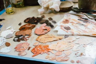 A variety of oil paints are mixed on a palette with shades ranging from white and beige to browns and pinks. The scene includes paint blobs on a mixing surface, some brushes, and a blurred background with a bottle and other materials. Used paper towels smeared with paint add to the creative clutter.