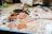 A variety of oil paints are mixed on a palette with shades ranging from white and beige to browns and pinks. The scene includes paint blobs on a mixing surface, some brushes, and a blurred background with a bottle and other materials. Used paper towels smeared with paint add to the creative clutter.