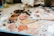 A variety of oil paints are mixed on a palette with shades ranging from white and beige to browns and pinks. The scene includes paint blobs on a mixing surface, some brushes, and a blurred background with a bottle and other materials. Used paper towels smeared with paint add to the creative clutter.