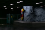 A dimly lit underground area featuring a large, colorful letter 'S' mounted on a rugged rock-like wall. The setting has industrial elements such as metal fencing and fluorescent lighting.