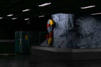 A dimly lit underground area featuring a large, colorful letter 'S' mounted on a rugged rock-like wall. The setting has industrial elements such as metal fencing and fluorescent lighting.
