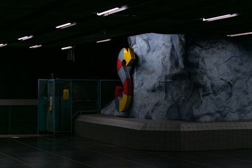 A dimly lit underground area featuring a large, colorful letter 'S' mounted on a rugged rock-like wall. The setting has industrial elements such as metal fencing and fluorescent lighting.