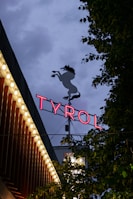 A neon sign with the word 'TYROL' illuminated in pink against a dusky sky, accompanied by a silhouette of a prancing horse above. Surrounding the sign, there are trees with dark green leaves and a building adorned with small round lights along its roofline, creating a vibrant and lively atmosphere.