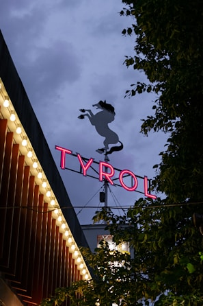 A neon sign with the word 'TYROL' illuminated in pink against a dusky sky, accompanied by a silhouette of a prancing horse above. Surrounding the sign, there are trees with dark green leaves and a building adorned with small round lights along its roofline, creating a vibrant and lively atmosphere.