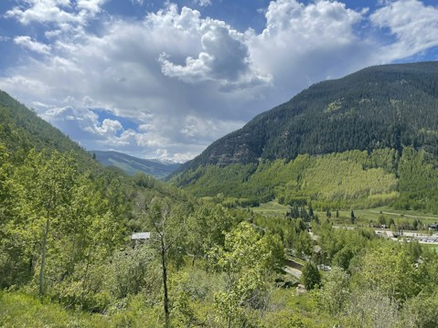 A lush, green valley with dense forests covering the rolling hills and mountains in the background. The sky is partly cloudy with patches of blue visible. A small building is nestled among the trees, blending into the verdant landscape.