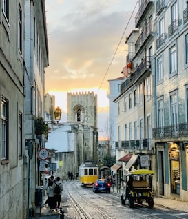 A narrow European street scene with cobblestone pavement features an iconic yellow tram traveling towards a historic stone building under a warm, glowing sunset. Pedestrians walk along the sidewalk, while parked vehicles line the street. The buildings are adorned with balconies and windows, showcasing traditional architecture.