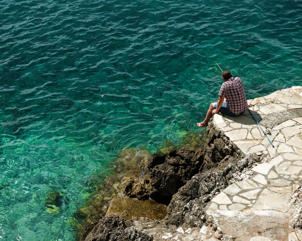 a man sitting on the edge of a cliff next to the ocean
