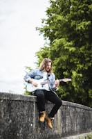A person with long hair is sitting on a stone wall playing an electric guitar. They are wearing a denim jacket, dark pants, and tan boots. Behind them, lush green trees create a natural backdrop.