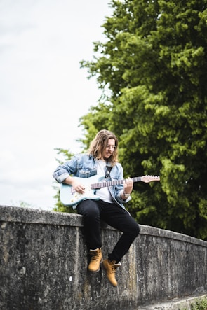 A person with long hair is sitting on a stone wall playing an electric guitar. They are wearing a denim jacket, dark pants, and tan boots. Behind them, lush green trees create a natural backdrop.