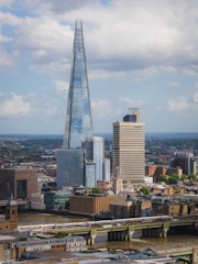 A cityscape featuring a tall, modern skyscraper with a sharp pointed top, surrounded by other buildings of varying architectural styles. A river flows in the foreground, crossed by a bridge carrying a train. The sky is partly cloudy.