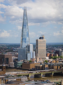 A cityscape featuring a tall, modern skyscraper with a sharp pointed top, surrounded by other buildings of varying architectural styles. A river flows in the foreground, crossed by a bridge carrying a train. The sky is partly cloudy.