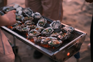 A grill filled with open oysters, which are garnished with colorful toppings, such as diced vegetables. The grill is set outdoors, possibly on a beach, with warm amber tones suggesting a sunset or evening setting. Several people are gathered around, engaged in the cooking process.