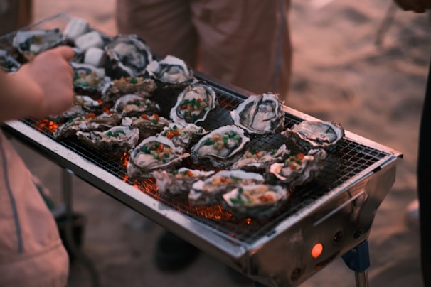 A grill filled with open oysters, which are garnished with colorful toppings, such as diced vegetables. The grill is set outdoors, possibly on a beach, with warm amber tones suggesting a sunset or evening setting. Several people are gathered around, engaged in the cooking process.