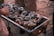 A grill filled with open oysters, which are garnished with colorful toppings, such as diced vegetables. The grill is set outdoors, possibly on a beach, with warm amber tones suggesting a sunset or evening setting. Several people are gathered around, engaged in the cooking process.