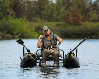 A person is sitting on a small inflatable raft in the middle of a calm body of water, holding a fishing rod. The individual is wearing a cap, camouflage safety vest, and outdoor attire appropriate for fishing. The background consists of lush greenery with trees and bushes.