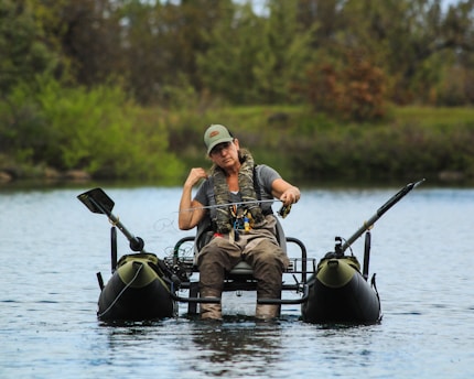 A person is sitting on a small inflatable raft in the middle of a calm body of water, holding a fishing rod. The individual is wearing a cap, camouflage safety vest, and outdoor attire appropriate for fishing. The background consists of lush greenery with trees and bushes.