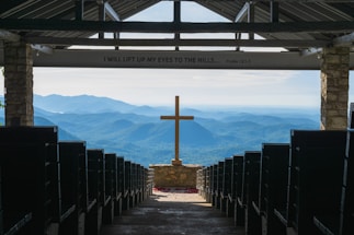 An open chapel setting with rows of pews leading to a prominent wooden cross at the center. Beyond the cross, the background features a stunning view of rolling blue mountains under a clear sky. The interior is framed by stone pillars, and above, an inscription reads 'I WILL LIFT UP MY EYES TO THE HILLS... Psalm 121:1.'
