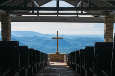 An open chapel setting with rows of pews leading to a prominent wooden cross at the center. Beyond the cross, the background features a stunning view of rolling blue mountains under a clear sky. The interior is framed by stone pillars, and above, an inscription reads 'I WILL LIFT UP MY EYES TO THE HILLS... Psalm 121:1.'
