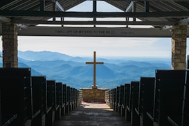 An open chapel setting with rows of pews leading to a prominent wooden cross at the center. Beyond the cross, the background features a stunning view of rolling blue mountains under a clear sky. The interior is framed by stone pillars, and above, an inscription reads 'I WILL LIFT UP MY EYES TO THE HILLS... Psalm 121:1.'