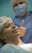 A woman lying down wearing a surgical cap and gown, smiling gently. Behind her stands a person in scrubs, mask, and glasses giving a thumbs-up gesture.
