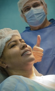 A woman lying down wearing a surgical cap and gown, smiling gently. Behind her stands a person in scrubs, mask, and glasses giving a thumbs-up gesture.