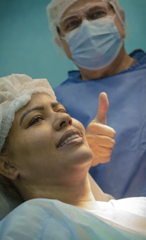 A woman lying down wearing a surgical cap and gown, smiling gently. Behind her stands a person in scrubs, mask, and glasses giving a thumbs-up gesture.