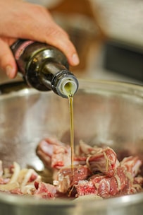 A hand is pouring oil from a dark bottle into a bowl containing raw meat pieces.