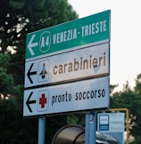 A road sign with directions to Venezia-Trieste on the A4 highway, along with signs indicating directions for a carabinieri station and an emergency medical service. The background shows some blurred greenery.