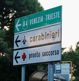 A road sign with directions to Venezia-Trieste on the A4 highway, along with signs indicating directions for a carabinieri station and an emergency medical service. The background shows some blurred greenery.