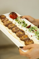 A close-up of a rectangular white plate being held, displaying a row of grilled meat rolls on a creamy sauce base. Garnished with green herbs, pink peppercorns, and sliced onions, alongside a charred tomato and a wedge of lemon. The hands with dark nail polish are visible holding the plate.